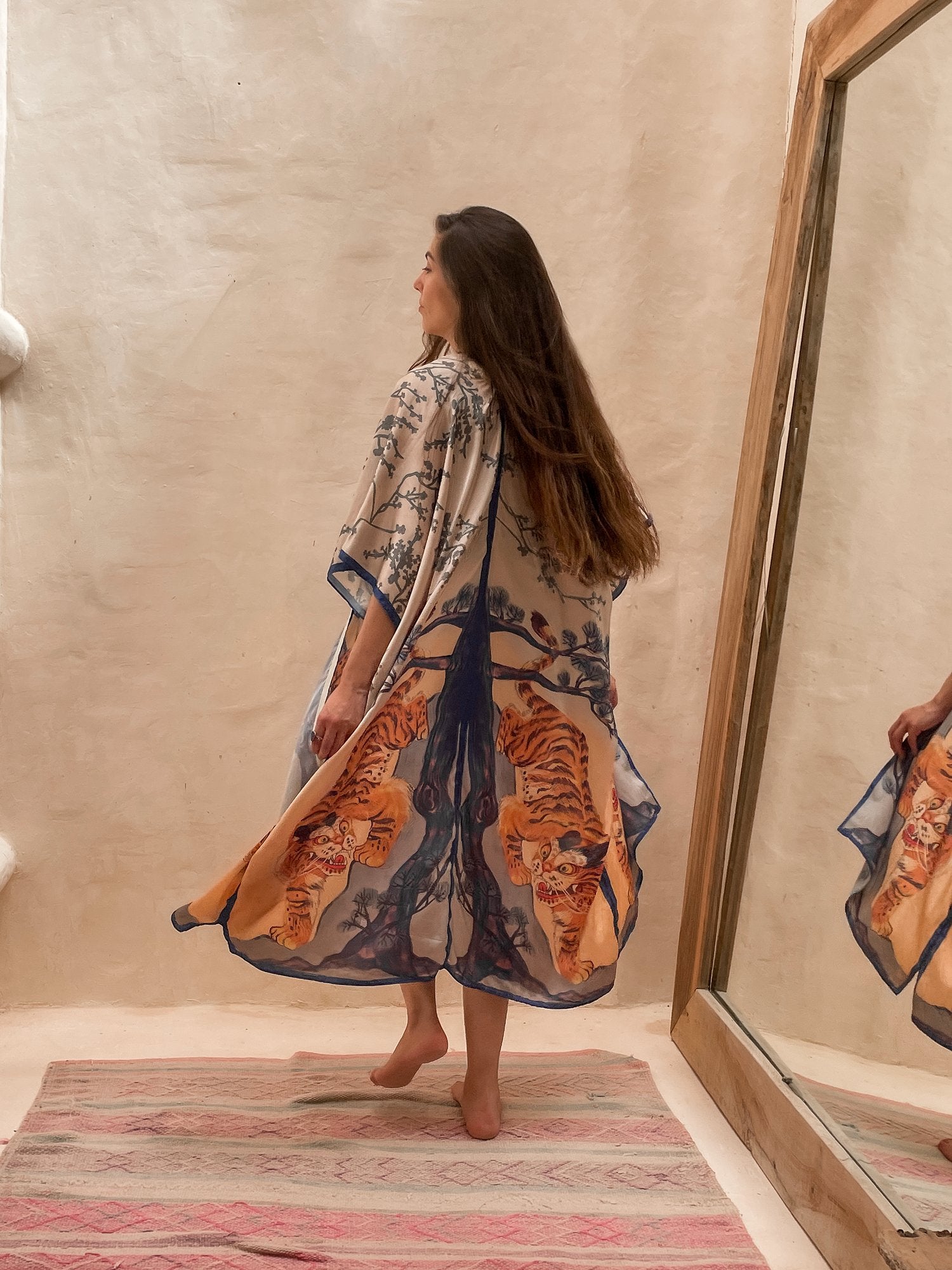 Model wearing the Kimono Bengal Tiger. She is towards the wall with a large mirror next to her. The design features a mirrored design of a large tree, nature details, and the bengal tiger.