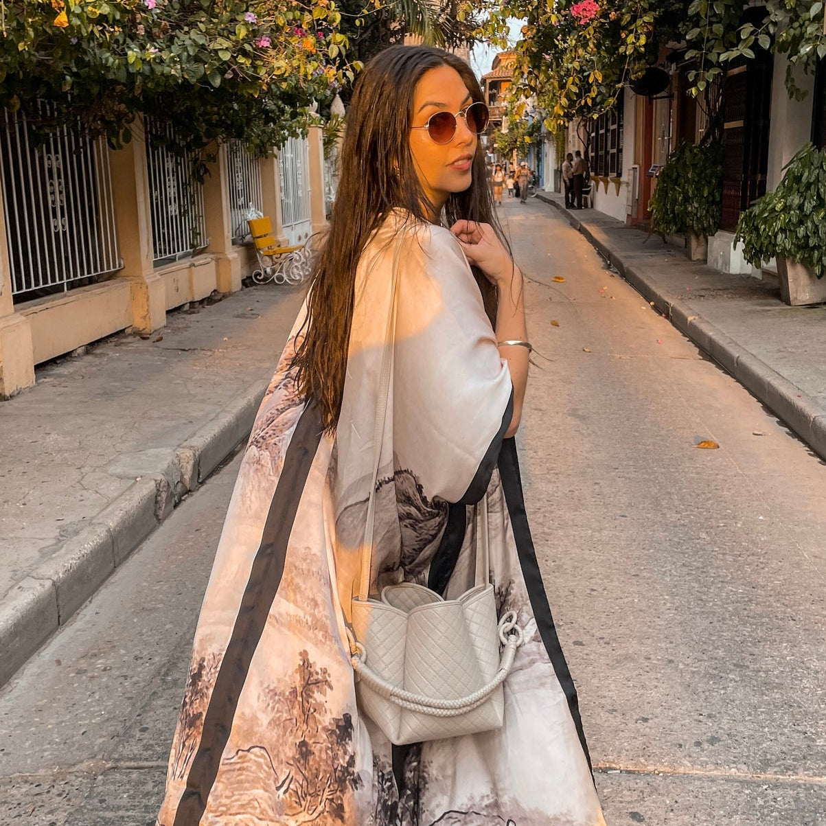 Model is wearing the Osaka Kimono by PLISSÉ. She is modeling the back of the kimono paired with the Baletti Tulip Bag in Off White. The pattern of the kimono is a neutral toned mountainscape.