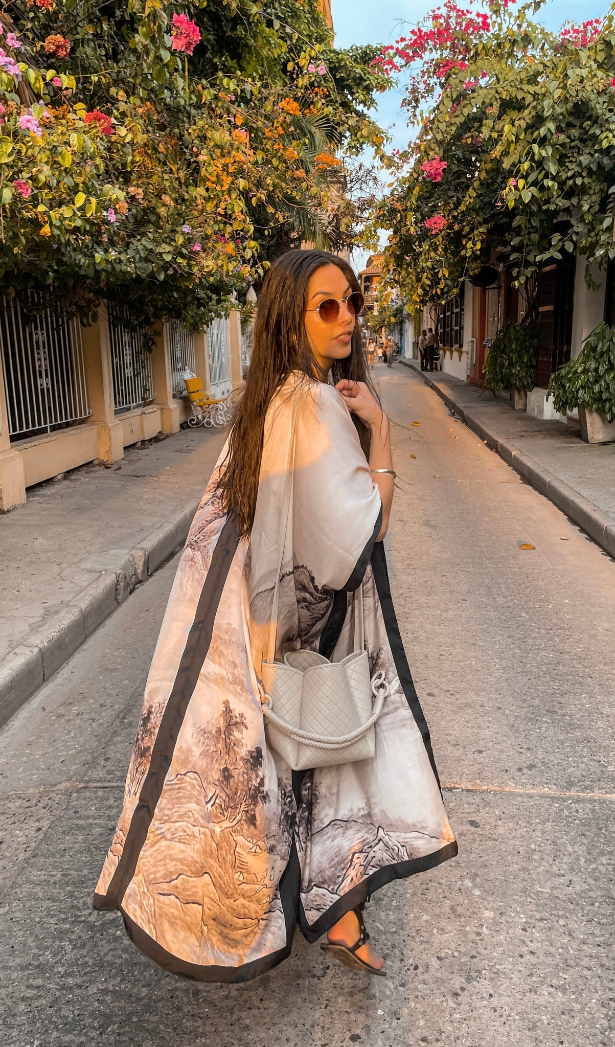 Model is wearing the Osaka Kimono by PLISSÉ. She is modeling the back of the kimono paired with the Baletti Tulip Bag in Off White. The pattern of the kimono is a neutral toned mountainscape.