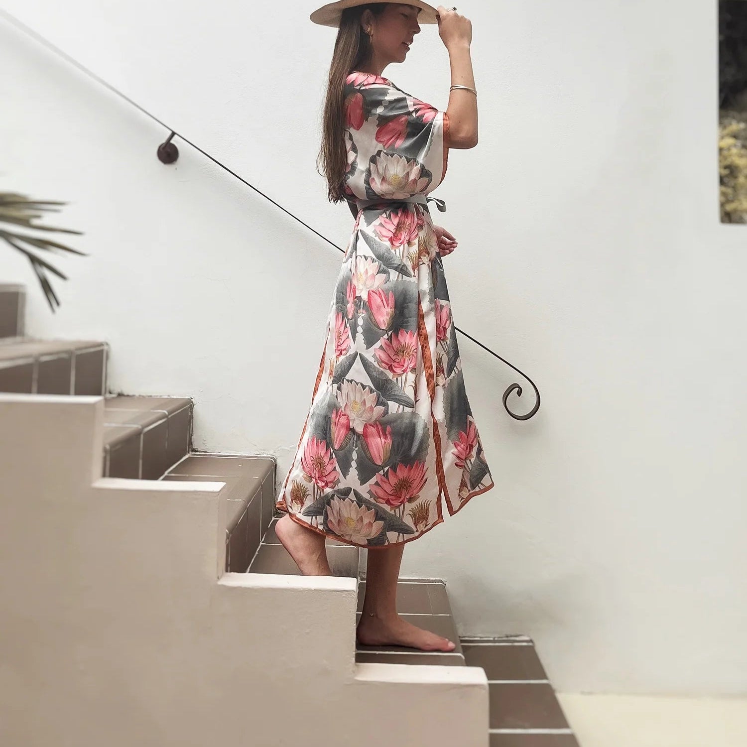 Model standing on white staircase, with her side towards us wearing the Lotus Kimono. She is modeling the Vibrant Style along with a natural colored hat, barefoot, demonstrating it is a good piece to lounge around the house as well.