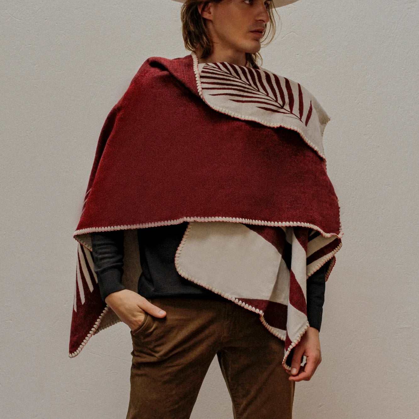 Man wearing Red Natura Poncho, standing on plain white wall. 