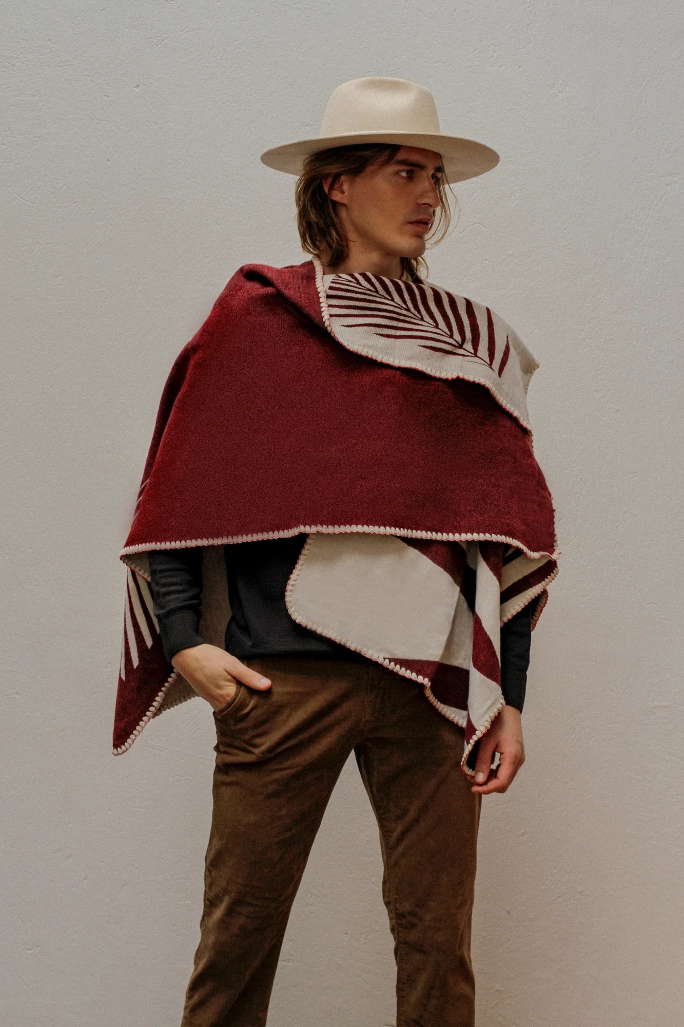 Man wearing Red Natura Poncho, standing on plain white wall. 