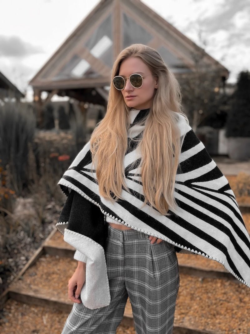 Natura Poncho by PLISSÉ. Model is wearing the poncho outside paired with grey plaid pants and sunglasses. She is standing in front of a greenhouse on a cloudy day, looking to floor.