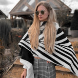Natura Poncho by PLISSÉ. Model is wearing the poncho outside paired with grey plaid pants and sunglasses. She is standing in front of a greenhouse on a cloudy day, looking to floor.