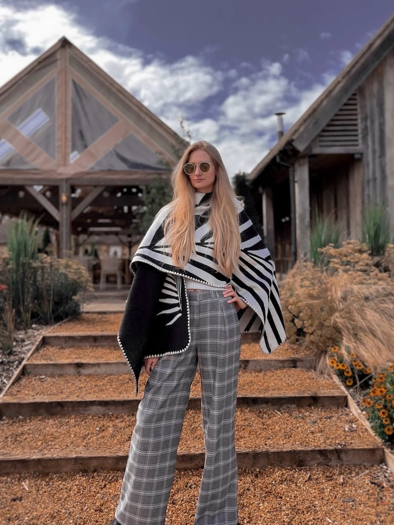 Natura Poncho by PLISSÉ. Model is wearing the poncho outside paired with grey plaid pants and sunglasses. She is standing in front of a greenhouse on a cloudy day, looking at camera.