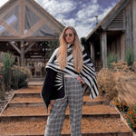 Natura Poncho by PLISSÉ. Model is wearing the poncho outside paired with grey plaid pants and sunglasses. She is standing in front of a greenhouse on a cloudy day, looking at camera.