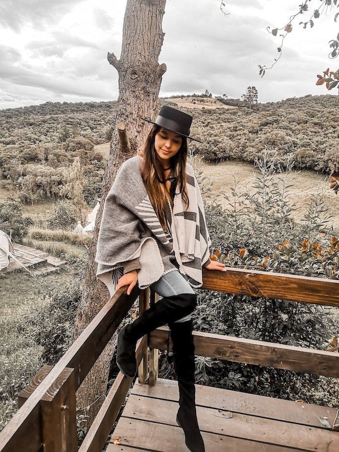 Paloma wearing grey natura poncho sitting on wooden rail. There is a view of a mountain and lots of natural plants.