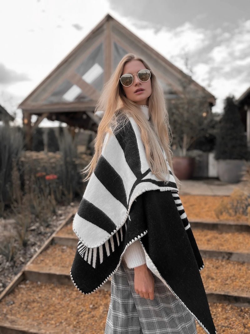 Natura Poncho by PLISSÉ. Model is wearing the poncho outside paired with grey plaid pants and sunglasses. She is standing in front of a greenhouse on a cloudy day, looking at camera.