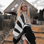 Natura Poncho by PLISSÉ. Model is wearing the poncho outside paired with grey plaid pants and sunglasses. She is standing in front of a greenhouse on a cloudy day, looking at camera.