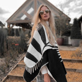 Natura Poncho by PLISSÉ. Model is wearing the poncho outside paired with grey plaid pants and sunglasses. She is standing in front of a greenhouse on a cloudy day, looking at camera.