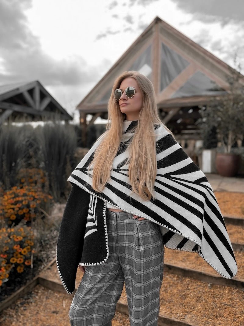 Natura Poncho by PLISSÉ. Model is wearing the poncho outside paired with grey plaid pants and sunglasses. She is standing in front of a greenhouse on a cloudy day, looking at away.