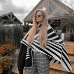 Natura Poncho by PLISSÉ. Model is wearing the poncho outside paired with grey plaid pants and sunglasses. She is standing in front of a greenhouse on a cloudy day, looking at away.