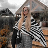 Natura Poncho by PLISSÉ. Model is wearing the poncho outside paired with grey plaid pants and sunglasses. She is standing in front of a greenhouse on a cloudy day, looking at away.
