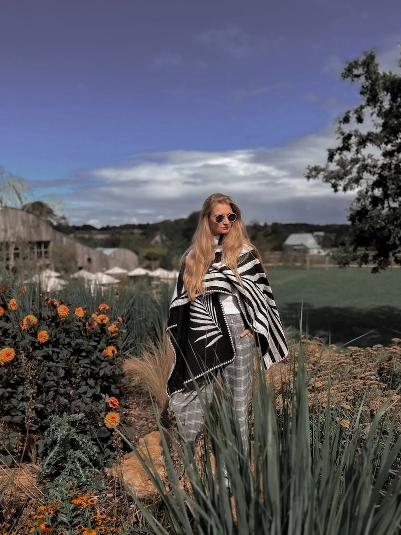 Natura Poncho by PLISSÉ. Model is wearing the poncho outside paired with grey plaid pants and sunglasses. She is standing on plants on a cloudy day, looking at camera.