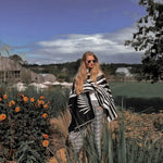 Natura Poncho by PLISSÉ. Model is wearing the poncho outside paired with grey plaid pants and sunglasses. She is standing on plants on a cloudy day, looking at camera.