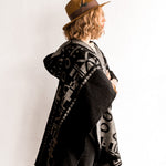 Side view of the woman wearing the OX Black and Grey Poncho without the hood. She paired the poncho with a brown hat, light brown top, and black jeans.