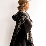 Side view of the woman wearing the OX Black and Grey Poncho without the hood. She paired the poncho with a brown hat, light brown top, and black jeans.