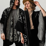 Man wearing the OX Black and Grey Poncho with Hood. He paired it with a grey top and black jeans. He is over a plain grey background. The women next to him wearing the same XO poncho. She paired the poncho with a brown hat, light brown top, and black jeans.