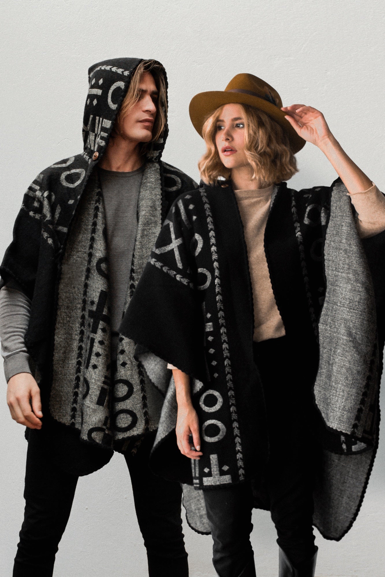 Man wearing the OX Black and Grey Poncho with Hood. He paired it with a grey top and black jeans. He is over a plain grey background. The women next to him wearing the same XO poncho. She paired the poncho with a brown hat, light brown top, and black jeans.