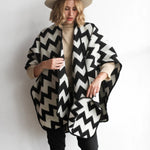 Woman modeling the picos ponchos. The pattern features a zig zag pattern across the whole poncho. She wore it with a light brown hat, cream top, and black jeans.