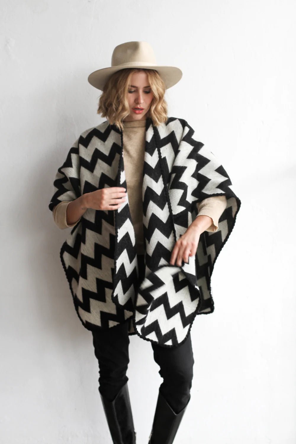 Woman modeling the picos ponchos. The pattern features a zig zag pattern across the whole poncho. She wore it with a light brown hat, cream top, and black jeans.