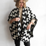 Woman modeling the picos ponchos. The pattern features a zig zag pattern across the whole poncho. She wore it with a light brown hat, cream top, and black jeans.