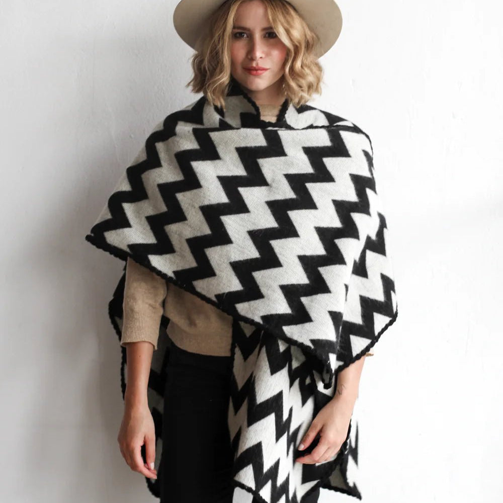 Woman modeling the picos ponchos. The pattern features a zig zag pattern across the whole poncho. She wore it with a light brown hat, cream top, and black jeans.