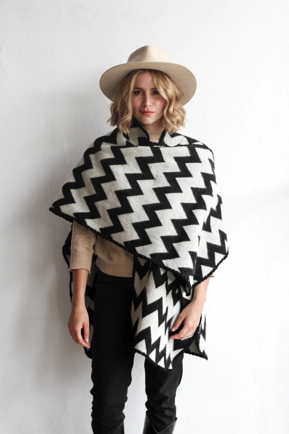 Woman modeling the picos ponchos. The pattern features a zig zag pattern across the whole poncho. She wore it with a light brown hat, cream top, and black jeans.