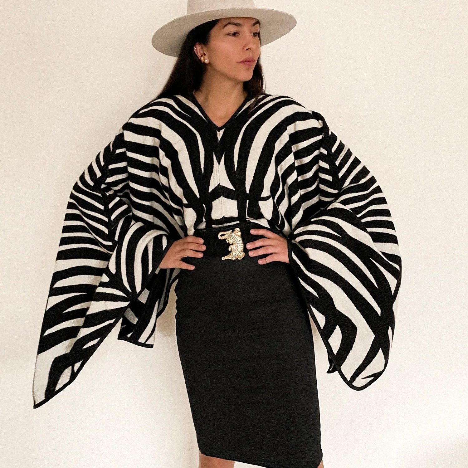 Model wearing the Africa Poncho with a belt and a black pencil skirt. She is showing us the front of the poncho. The Africa poncho features a zebra pattern. The poncho is paired with a white hat. Model is standing on a white wall.