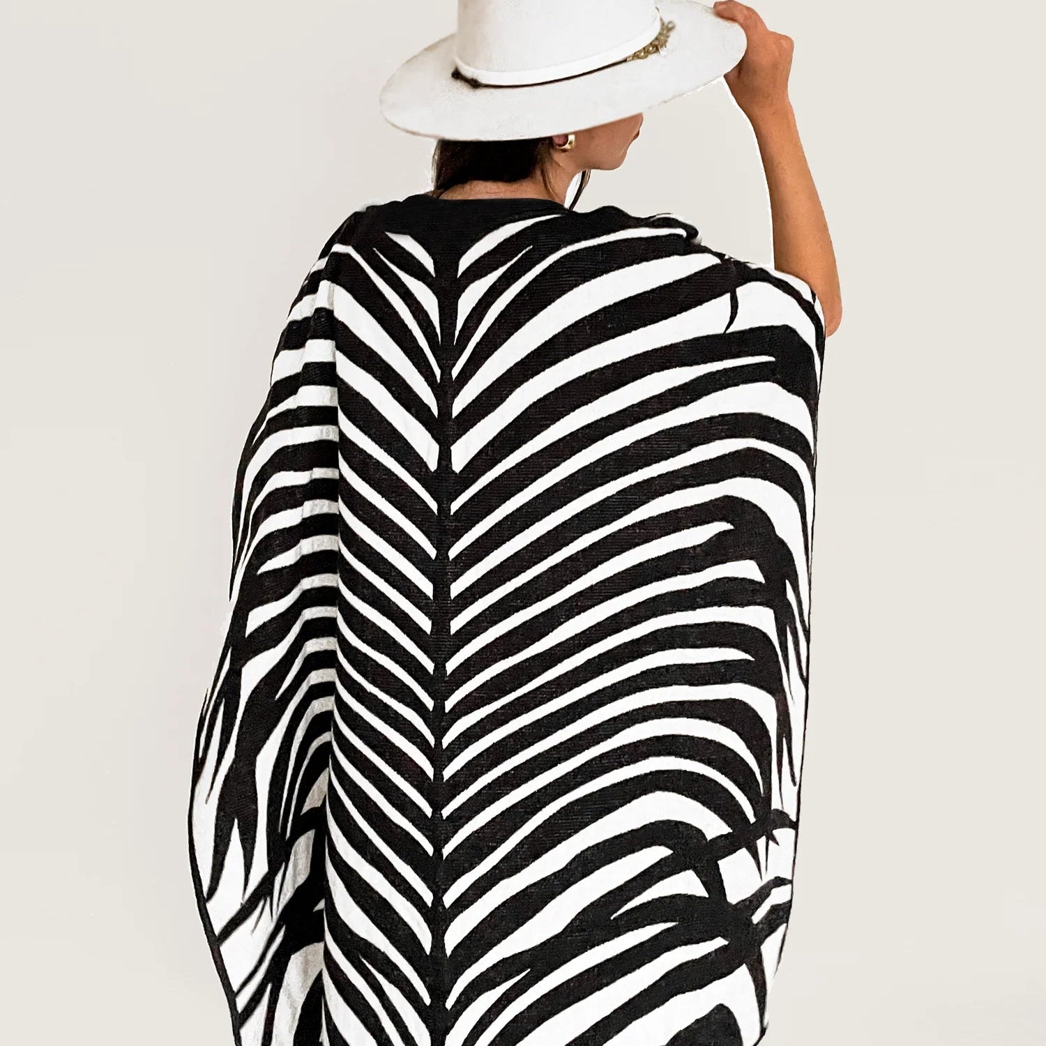 Model wearing the Africa Poncho with black jeans. She is showing us the back of the poncho. The Africa poncho features a zebra pattern. The poncho is paired with a white hat. Model is standing on a white wall.