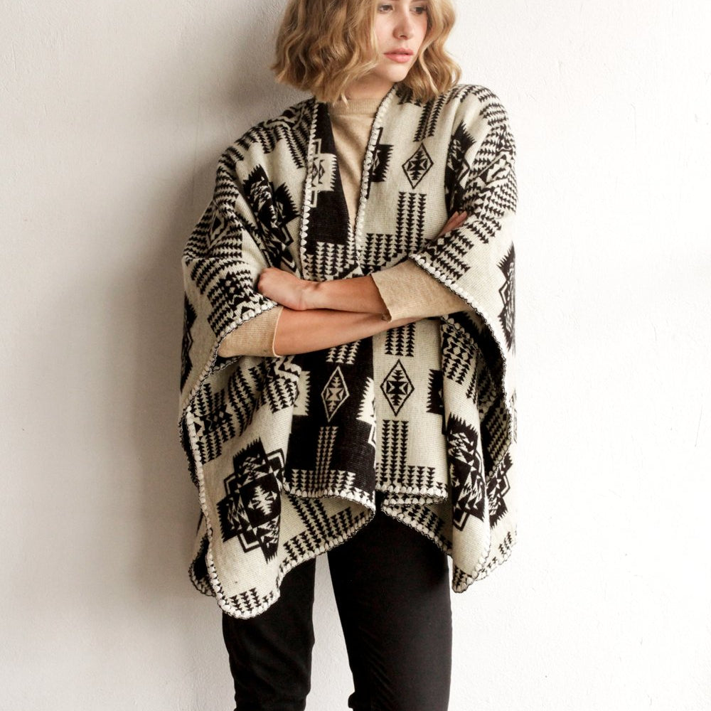 She is wearing the black sierra poncho. Poncho features a geometric design with tones darker on one side. She is standing on a plain white wall.