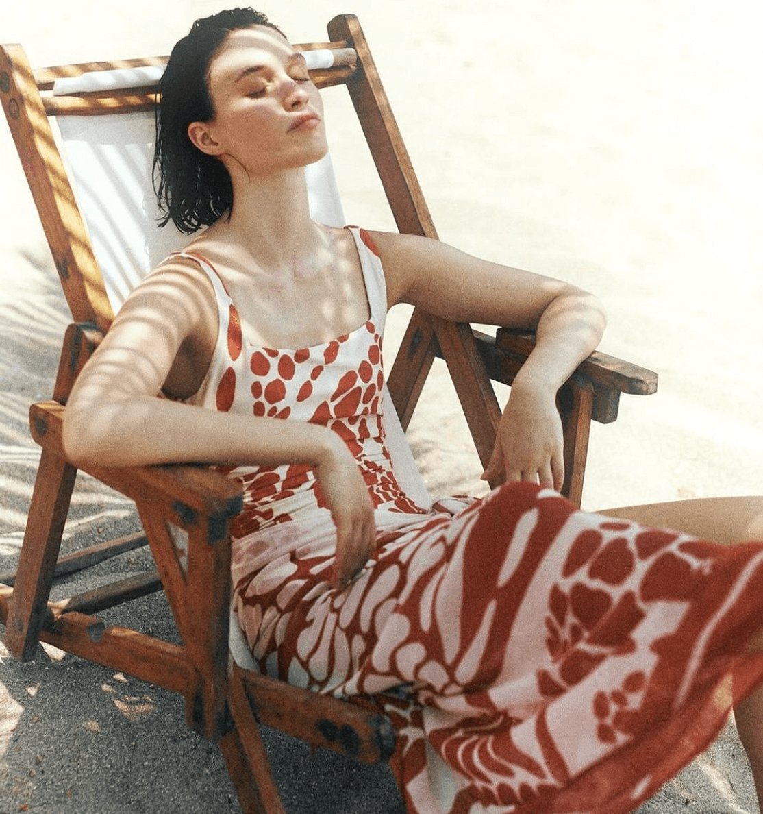 Model laying down on beach chair. She is at the beach relaxing and posing with eyes closed. She is wearing the Sole One Piece Swimsuit in Orange Mare with its matching sarong. 