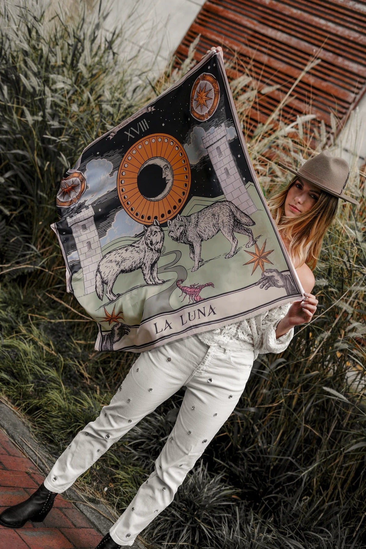 Model holding up the Zodiac Moon Scarf. Scene has brick flooring, plants behind her.
