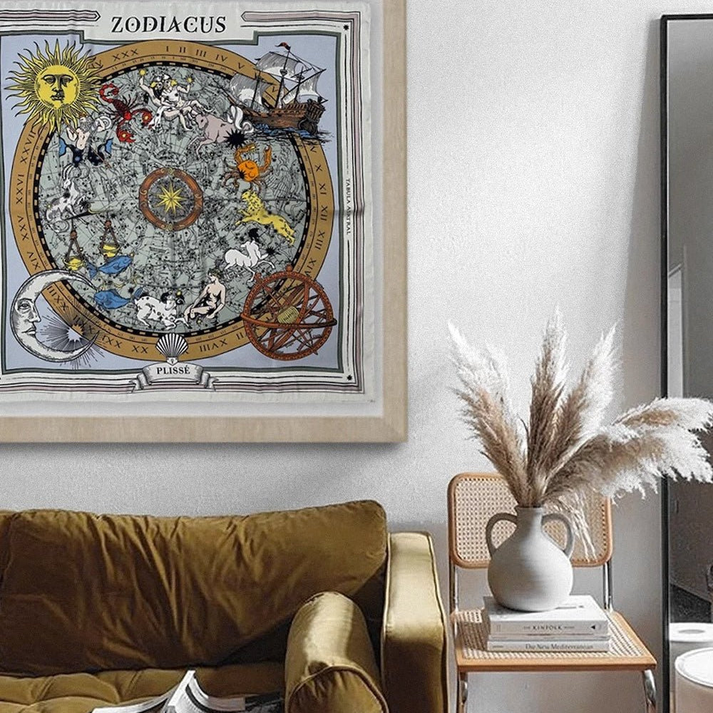 Brown zodiac scarf displayed in a frame over a couch, and natural chair with books and stone vase. 