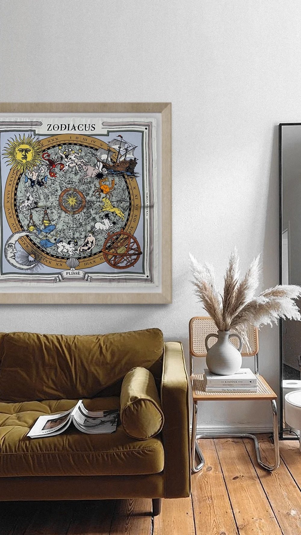 Brown zodiac scarf displayed in a frame over a couch, and natural chair with books and stone vase. 