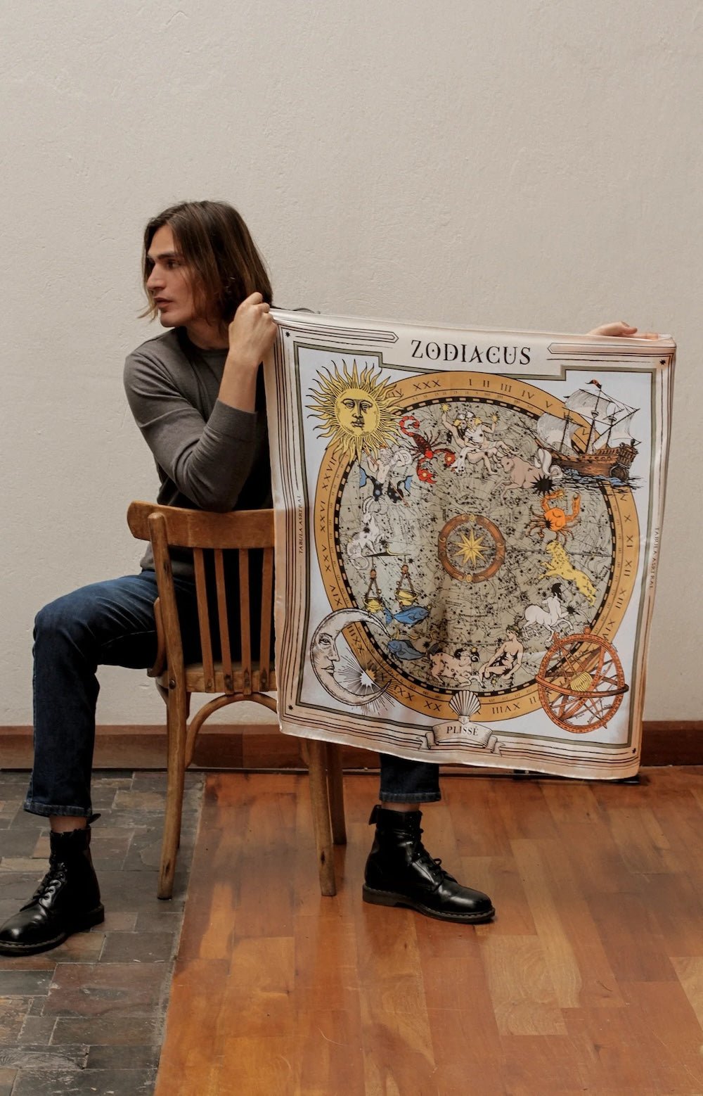 Man holding up brown zodiac scarf while sitting on chair backwards