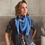 Model is wearing Zodiac Scarf for Aquarius wrapped around neck with grey top and jeans. She is sitting near concrete wall with cactus behind her. 