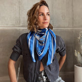 Model is wearing Zodiac Scarf for Aquarius wrapped around neck with grey top and jeans. She is sitting near concrete wall with cactus behind her. 