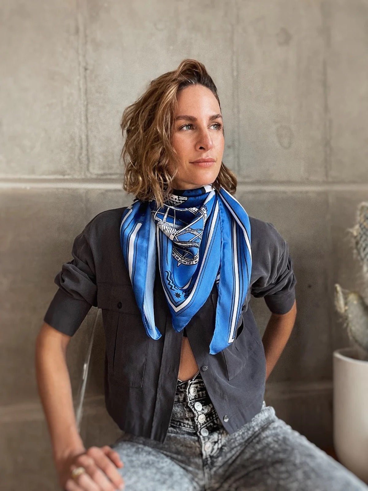 Model is wearing Zodiac Scarf for Aquarius wrapped around neck with grey top and jeans. She is sitting near concrete wall with cactus behind her. 