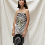 Model wearing Zodiac Scarf for Cancer wrapped on as a strapless top, paired with white jeans and grey hat. She is standing on a plain white background. 