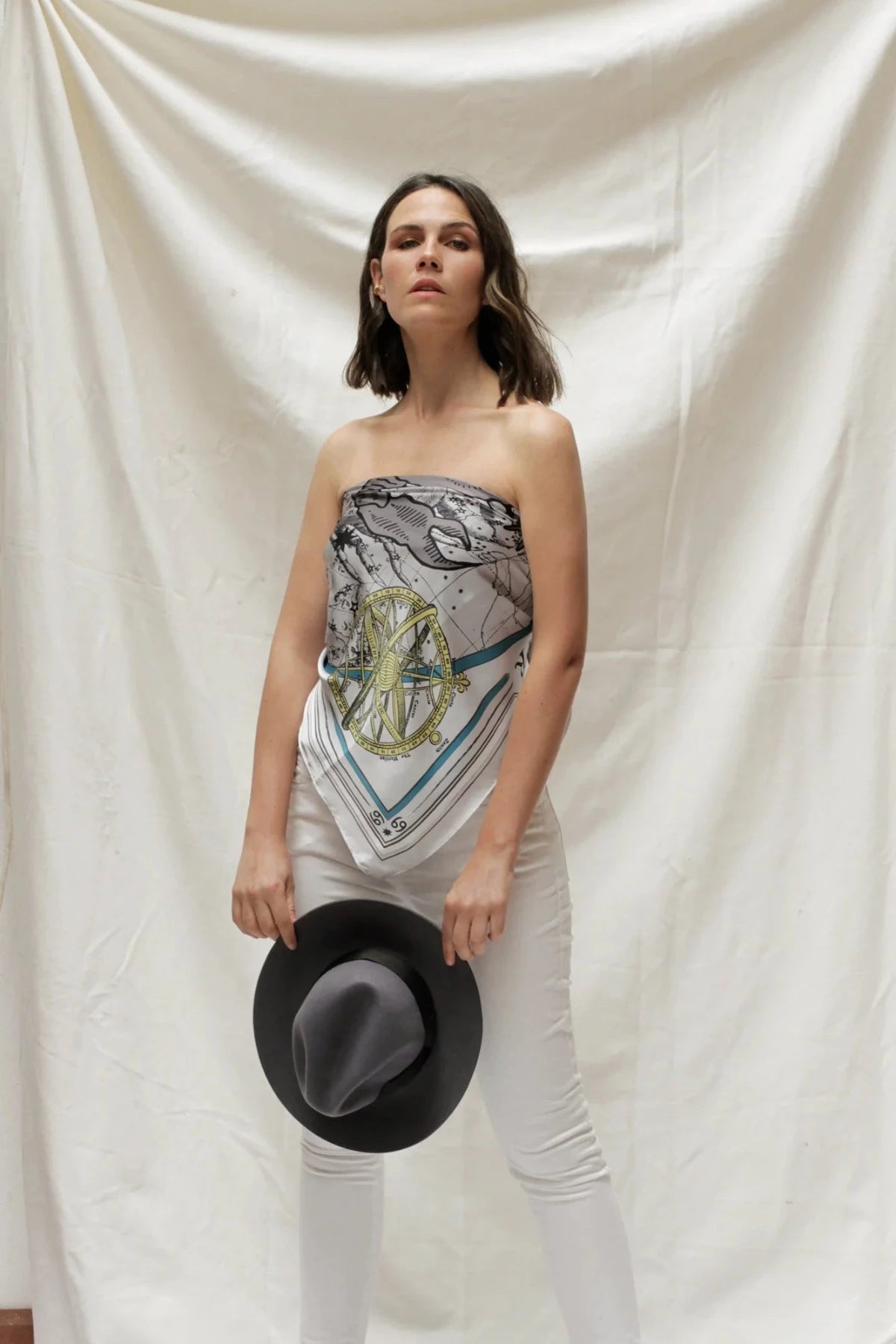 Model wearing Zodiac Scarf for Cancer wrapped on as a strapless top, paired with white jeans and grey hat. She is standing on a plain white background. 