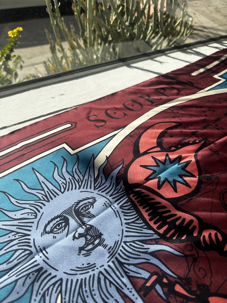 Close up of burgundy scorpio scarf by Plissé. Image of a scorpion, with sun and moon on corners. The scarf is pictured laid out on window of EVAMAIA.